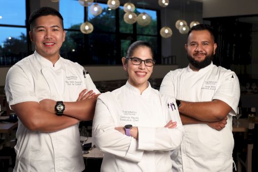 Three chefs wearing white uniforms stand with arms crossed, smiling in a well-lit restaurant setting.