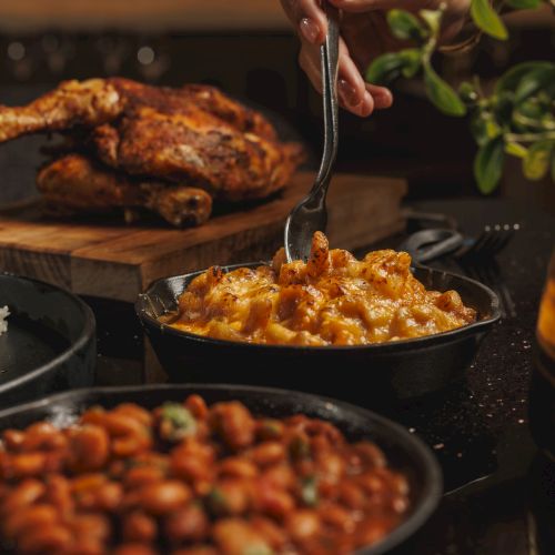 A hand scoops cheesy, spicy sauce from a skillet while grilled meat, corn, and veggies cook on a rustic grill.