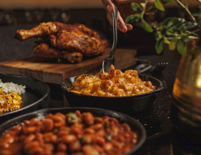 A hand scoops cheesy, spicy sauce from a skillet while grilled meat, corn, and veggies cook on a rustic grill.
