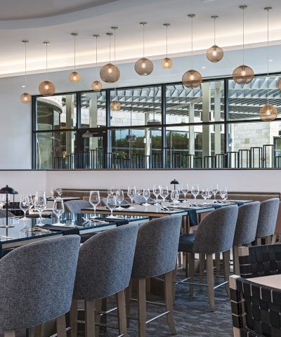 A modern, elegant restaurant with many gray upholstered chairs, long tables set with glassware and cutlery, and a row of hanging pendant lights above, creating a stylish dining space.