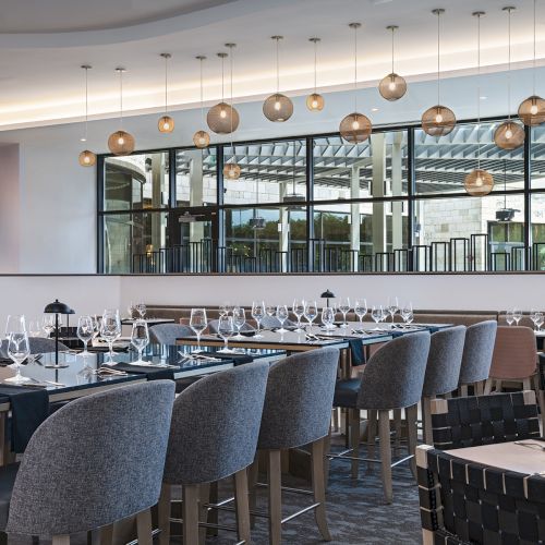 A modern, elegant restaurant with many gray upholstered chairs, long tables set with glassware and cutlery, and a row of hanging pendant lights above, creating a stylish dining space.
