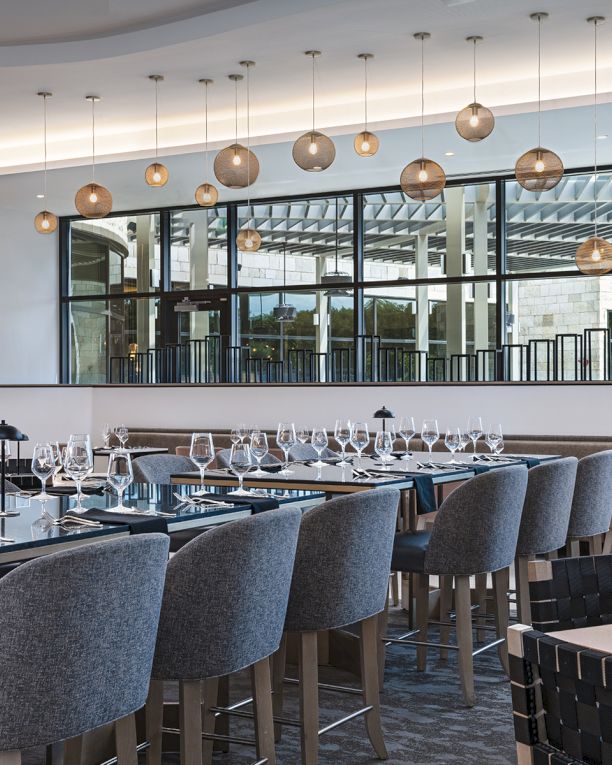 A modern, elegant restaurant with many gray upholstered chairs, long tables set with glassware and cutlery, and a row of hanging pendant lights above, creating a stylish dining space.