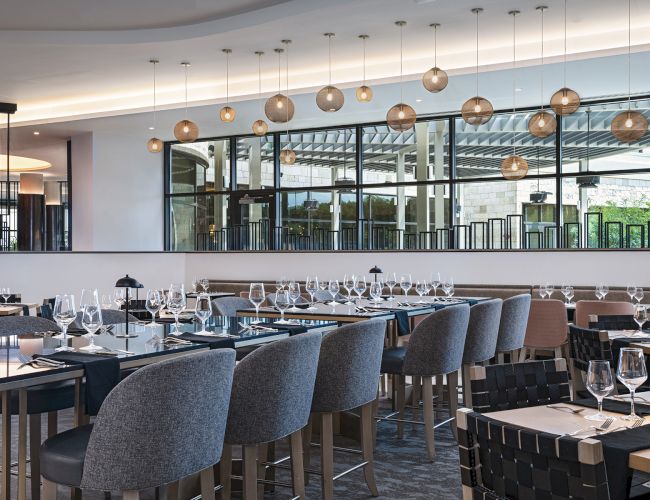A modern, elegant restaurant with many gray upholstered chairs, long tables set with glassware and cutlery, and a row of hanging pendant lights above, creating a stylish dining space.