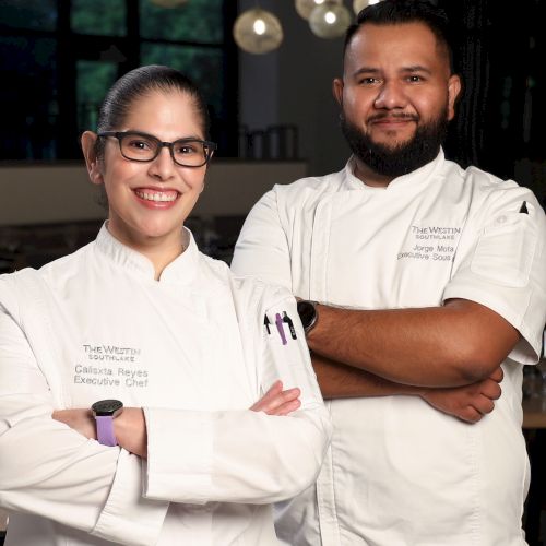 Two chefs in white uniforms pose together&mdash;female chef with glasses and a bearded male executive chef in a hotel kitchen.
