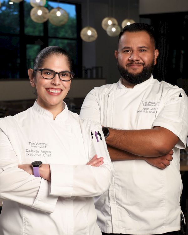 Two chefs in white uniforms pose together&mdash;female chef with glasses and a bearded male executive chef in a hotel kitchen.