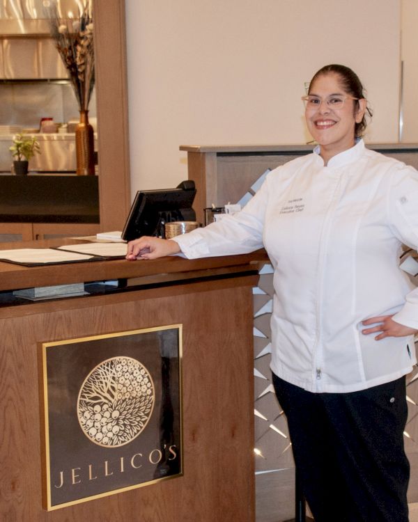 A chef in a white uniform stands at a restaurant reception desk with a 