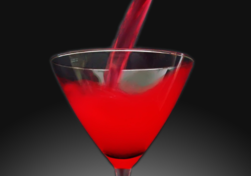 A red liquid being poured into a martini glass, with red roses placed at the bottom of the glass on a gradient gray background.