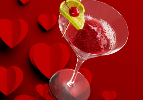 A red cocktail in a martini glass with a lime and cherry garnish, set against a background of red hearts.