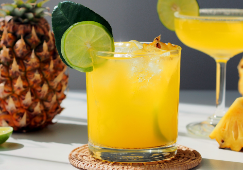 A cocktail with lime and pineapple garnishes, a whole pineapple, two drinks, and lime wedges are artfully arranged on a sunlit table.