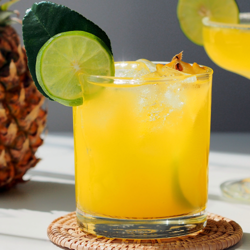 A cocktail with lime and pineapple garnishes, a whole pineapple, two drinks, and lime wedges are artfully arranged on a sunlit table.