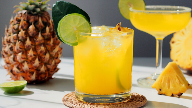 A cocktail with lime and pineapple garnishes, a whole pineapple, two drinks, and lime wedges are artfully arranged on a sunlit table.