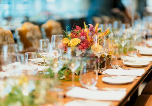 A long, elegantly set dining table with crystal glasses, white plates, napkins, and a vibrant floral centerpiece.