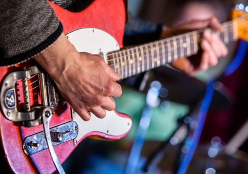 A person is playing a red electric guitar, focusing on the guitar's neck and body, with blurred background elements.