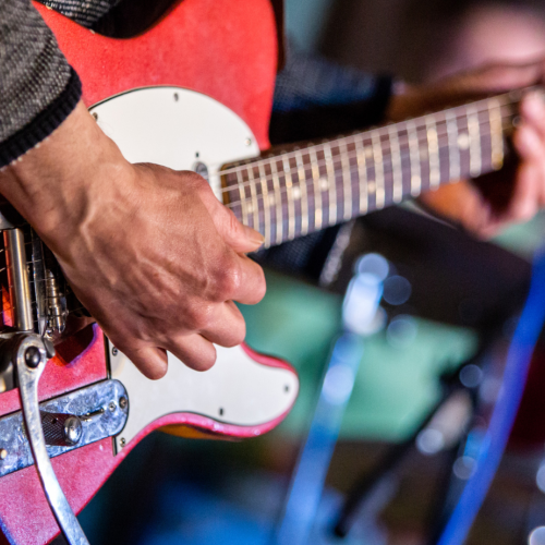 A person is playing a red electric guitar, focusing on the guitar's neck and body, with blurred background elements.