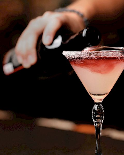 A person is pouring a dark liquid into a fancy cocktail glass with a salted rim, which has a light pink cocktail.