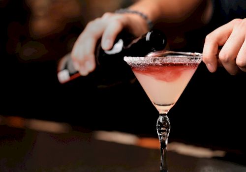 A person is pouring a dark liquid into a fancy cocktail glass with a salted rim, which has a light pink cocktail.