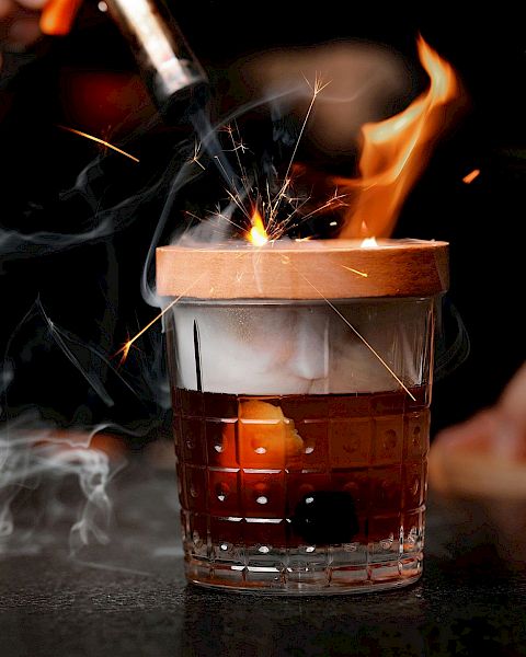 A person is using a blowtorch to ignite a cocktail, causing flames and sparks to appear around the glass, creating a dramatic presentation.