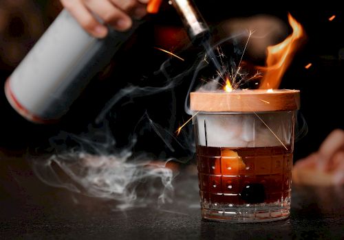 A person is using a blowtorch to ignite a cocktail, causing flames and sparks to appear around the glass, creating a dramatic presentation.