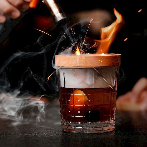 A person is using a blowtorch to ignite a cocktail, causing flames and sparks to appear around the glass, creating a dramatic presentation.