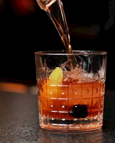 A drink being poured into a glass with a dark background. The glass contains ice, a lemon twist, and a cherry, possibly a cocktail.