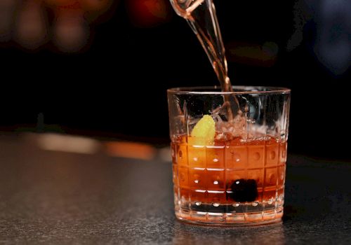 A drink being poured into a glass with a dark background. The glass contains ice, a lemon twist, and a cherry, possibly a cocktail.