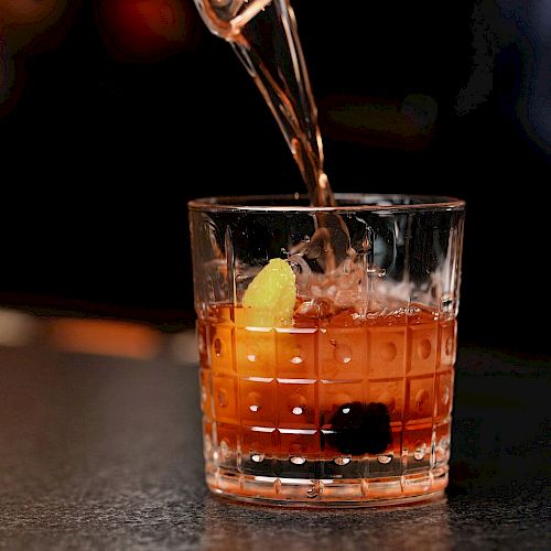 A drink being poured into a glass with a dark background. The glass contains ice, a lemon twist, and a cherry, possibly a cocktail.