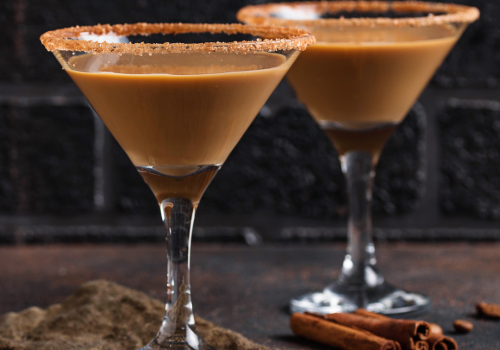 Two martini glasses filled with a creamy brown drink, sugar rimmed, beside cinnamon sticks on a dark surface.
