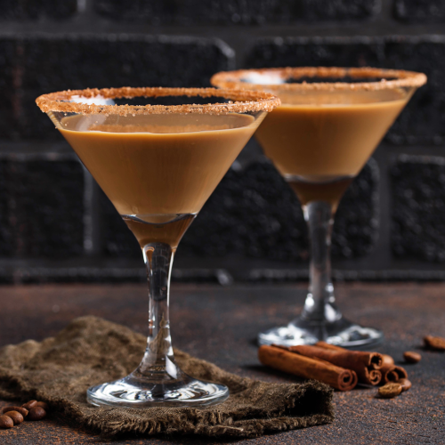 Two martini glasses filled with a creamy brown drink, sugar rimmed, beside cinnamon sticks on a dark surface.