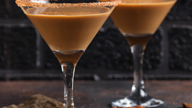 Two martini glasses filled with a creamy brown drink, sugar rimmed, beside cinnamon sticks on a dark surface.