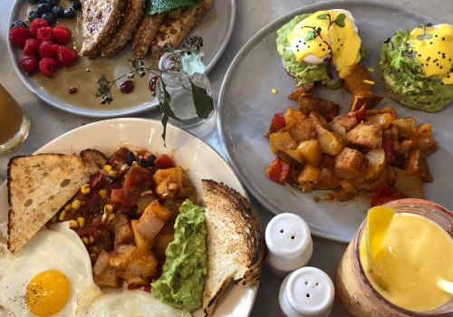 A breakfast spread with avocado toast, eggs Benedict, fried eggs, toast, potatoes, mixed berries, and drinks, including a smoothie and tea.