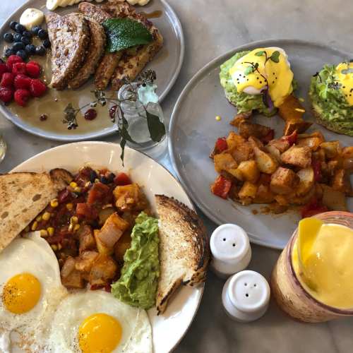 A breakfast spread with avocado toast, eggs Benedict, fried eggs, toast, potatoes, mixed berries, and drinks, including a smoothie and tea.