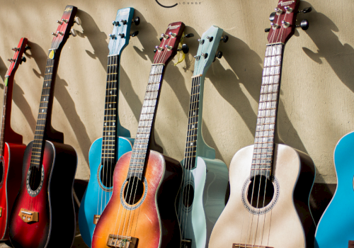 The image shows a row of colorful acoustic guitars leaned against a wall with a logo that reads 