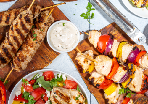 The image shows a variety of grilled dishes including kebabs, skewers with vegetables, sausage, and a grilled chicken-topped salad, with dip in the center.