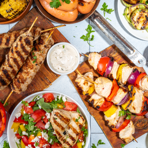 The image shows a variety of grilled dishes including kebabs, skewers with vegetables, sausage, and a grilled chicken-topped salad, with dip in the center.