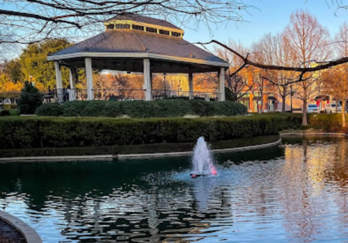 A serene park scene with a gazebo, surrounded by trees and bushes, overlooking a calm pond with a small fountain in the center.