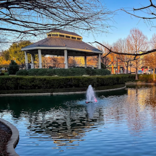 A serene park scene with a gazebo, surrounded by trees and bushes, overlooking a calm pond with a small fountain in the center.