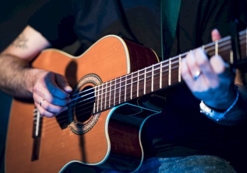 A person is playing an acoustic guitar, with their left hand on the fretboard and their right hand near the soundhole strumming or picking the strings.