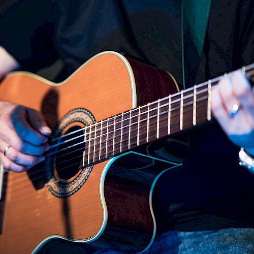 A person is playing an acoustic guitar, with their left hand on the fretboard and their right hand near the soundhole strumming or picking the strings.