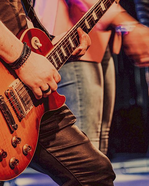 Close-up of a person playing an electric guitar on stage with other performers visible in the background.