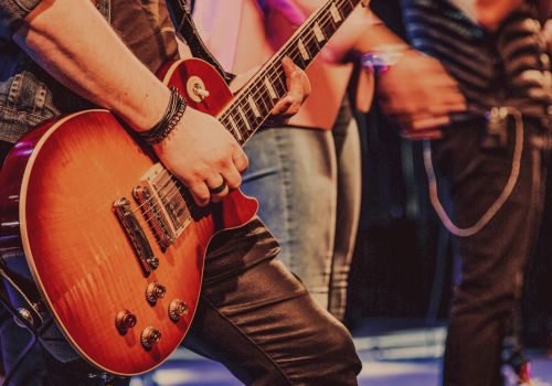 Close-up of a person playing an electric guitar on stage with other performers visible in the background.