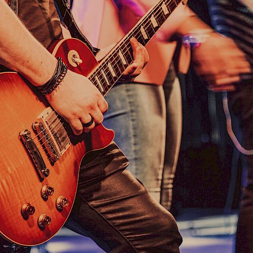 Close-up of a person playing an electric guitar on stage with other performers visible in the background.