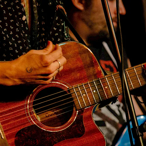 A musician strums a red acoustic guitar while a microphone stand sits nearby on a stage.