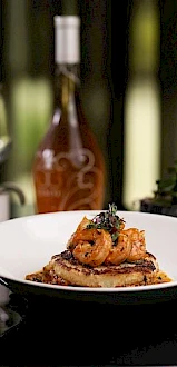 A dish with shrimp on top of a savory base is served on a white plate. Wine glasses and bottles are in the background, creating a dining ambiance.