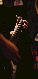 A person is playing an electric guitar in a dimly lit setting, focusing on the fretboard. The background appears dark with some blurred objects.