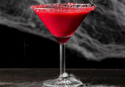 A red cocktail in a martini glass, rimmed with red sugar, surrounded by fake spiders and webs, against a spooky backdrop.