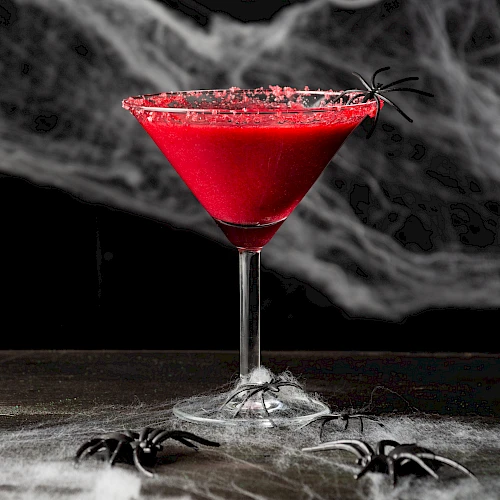 A red cocktail in a martini glass, rimmed with red sugar, surrounded by fake spiders and webs, against a spooky backdrop.