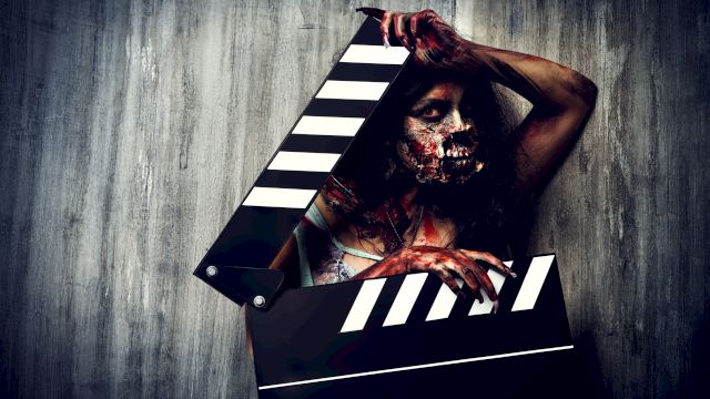 A person with zombie makeup holding a large clapperboard, standing against a textured wall, creating a dramatic horror theme.