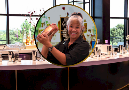 A bartender is shaking a cocktail mixer behind a bar counter with various beverages and glassware, indoors.