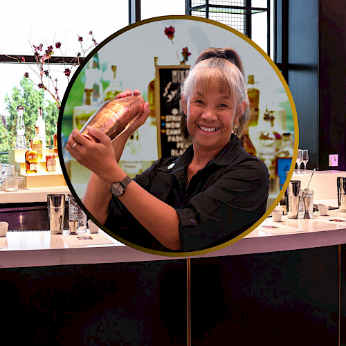 A bartender is shaking a cocktail mixer behind a bar counter with various beverages and glassware, indoors.