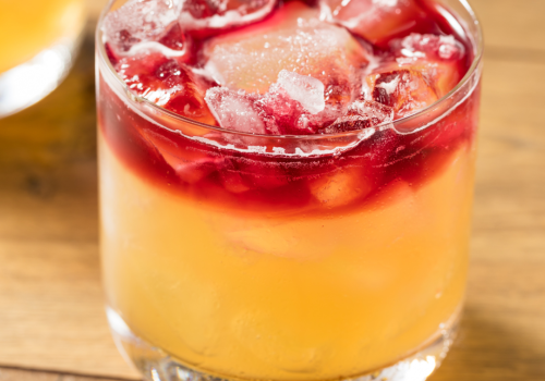 A cocktail with a yellow base, topped with red liquid and ice cubes in a short glass sitting on a wooden surface.